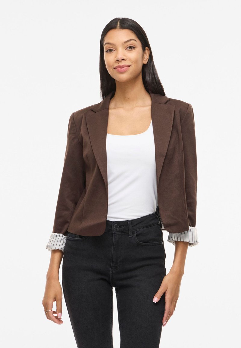 Femme aux longs cheveux foncés portant un blazer marron avec des revers rayés retroussés, un haut blanc et un jean noir taille haute, debout et souriante.