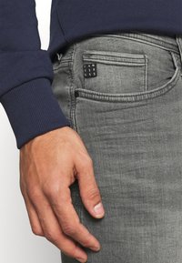 Šedé džíny z denimu s přiléhavým střihem, detail ruky opírající se o kapsu; obsahují malou černou štítek s bílým textem.