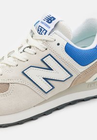 Tenisky New Balance 574 mají béžový svršek z semiše a síťoviny s modrými detaily, výrazné bílé logo "N" a texturovanou gumovou podrážku.