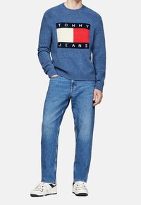 Blå stickad tröja med grafisk design, som har ett rött och vitt rektangel och texten "TOMMY JEANS". Kombinerad med ljusblå jeans och vita sneakers.