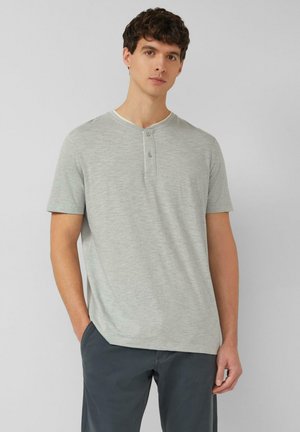 Junger Mann mit lockigem dunklem Haar, der ein hellgraues Henley-Shirt mit kurzen Ärmeln und dunkelgraue Hose trägt, stehend vor einem schlichten Hintergrund.