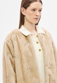 Casaco bege de pêlo sintético com detalhes florais, apresentando uma gola branca e botões dourados. Textura macia e corte oversized.