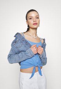 Giubbotto in denim blu chiaro indossato sopra un top a coste abbinato con un nodo frontale, abbinato a pantaloni bianchi a vita alta e accessori in perle.