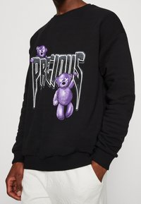 Felpa nera con una grafica di un orsacchiotto viola e il testo in grassetto che recita "PRECIOUS". Progettata con una vestibilità rilassata e polsini a coste.