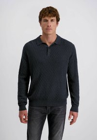 Man met kort krullend bruin haar, gekleed in een donker getextureerde polotrui met lange mouwen en vervaagde zwarte jeans, tegen een effen achtergrond.