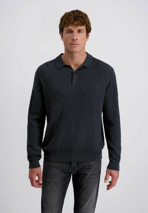 Man met kort krullend bruin haar, gekleed in een donker getextureerde polotrui met lange mouwen en vervaagde zwarte jeans, tegen een effen achtergrond.
