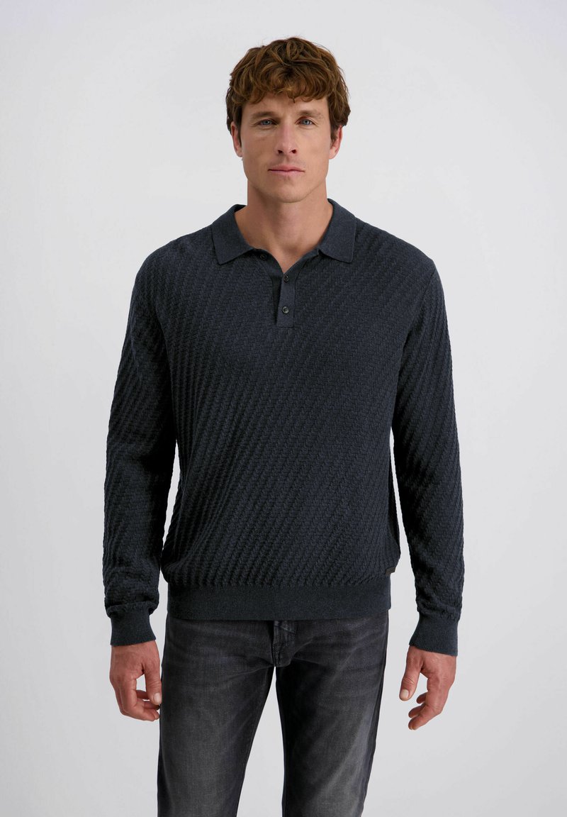 Man met kort krullend bruin haar, gekleed in een donker getextureerde polotrui met lange mouwen en vervaagde zwarte jeans, tegen een effen achtergrond.