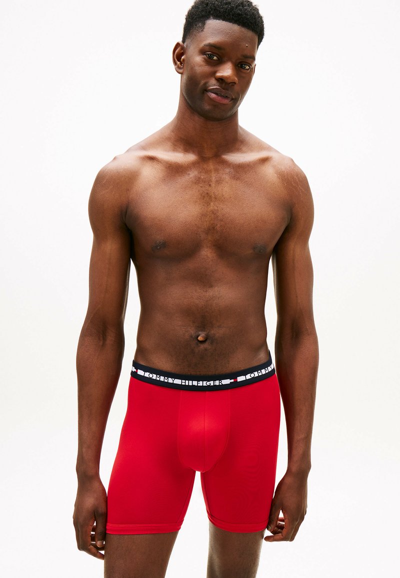 Boxer-briefs rouges en tissu lisse avec une taille noire arborant le logo "TOMMY HILFIGER" en blanc. Design ajusté avec une longueur mi-cuisse.
