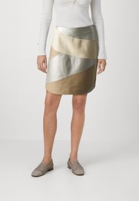 Metallisk mini skjørt med bølgende paneler i nyanser av beige, gull og sølv. Glatt tekstur med blank finish. Brukt sammen med lyse, vevde sko.