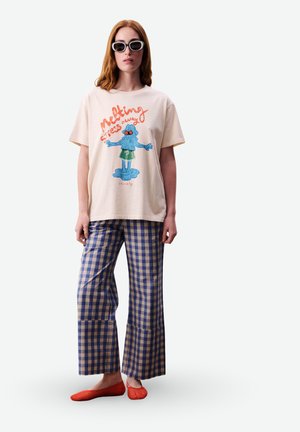 Femme portant un t-shirt crème avec un personnage bleu fondant et du texte, un pantalon large à carreaux bleu vichy, des chaussures rouges et des lunettes de soleil noires, debout.