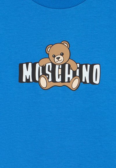 Μπλε βαμβακερό t-shirt με γραφικό μιας αρκουδάκι που κρατά τη λέξη "MOSCHINO" σε έντονη μαύρη και λευκή τυπογραφία.
