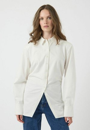 Vit långärmad skjorta med knappar och en strukturerad krage, med en framplacket och en rundad nederkant, i kombination med blå denimjeans.