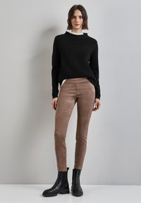 Maglione nero, tessuto leggero, scollatura alta, indossato sopra una camicia bianca; leggings in suede marrone con una texture liscia; stivali neri alla caviglia con suole spesse.