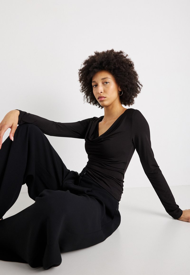 Črn dolg rokav s spuščenim ovratnikom, v kombinaciji s širokimi črnimi hlačami. Gladko blago z oprijetim silhueto in subtilnim sijajem.