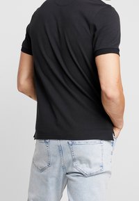 Man i svart kortärmad polo och ljusblå jeans, står med händerna i fickorna, med ryggen mot kameran.