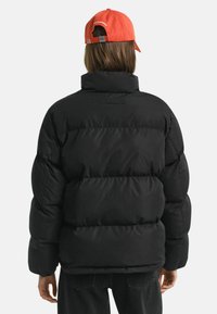 Zwarte pufferjas met hoge kraag en een gestikt ontwerp. Vergezeld door een oranje pet. Zichtbare stiksels en een relaxte pasvorm.