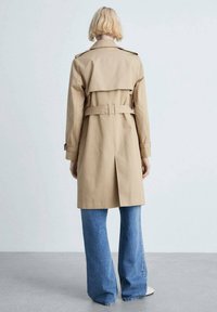 Trench-coat beige avec une taille ceinturée, une fente au dos et des épaulettes aux épaules, associé à un jean bleu évasé. Texture lisse et design classique.