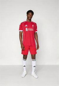 Röd fotbollströja med vita detaljer och logotyp, matchande röda shorts, vita strumpor med svarta ränder och vita fotbollsskor. Andningsbart tyg.