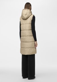 Beige prošívaná vesta s kapucí, sečleněným designem a rovnými liniemi. Nositá přes černý žebrovaný top a spárovaná s černými kalhotami.