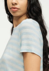 Νεαρό άτομο με μακριά σκούρα μαλλιά που φοράει ανοιχτό μπλε και λευκό ριγέ κοντομάνικη μπλούζα, δείχνοντας δαχτυλίδι στη μύτη στο αριστερό ρουθούνι.