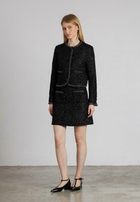 Chaqueta de tweed negra con un diseño ajustado, cuello redondo y dos bolsillos frontales, combinada con una falda mini a juego y zapatos negros.