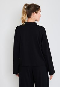 Czarna bluzka z długim rękawem o luźnym kroju, gładkiej fakturze i subtelnych detalach szwów. Posiada kołnierz typu mandarynka i opadające ramiona.