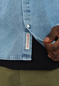 Lichtblauwe denimshirt met een geknoopte kraag, met een rode "REVOLUTION" label aan de zoom. De achtergrond bevat een zwart en olijfgroene laag.