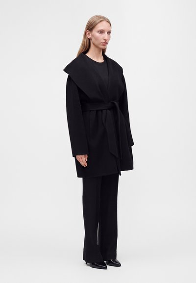Femme debout portant un manteau noir ceinturé avec un large col, pantalon noir et chaussures noires à bouts pointus sur un fond clair uni.