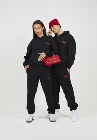 Μαύρες κουκούλες και φόρμες με κόκκινη κεντημένη λεπτομέρεια, σε συνδυασμό με μια κόκκινη τσάντα. Λευκά αθλητικά παπούτσια και ένα κόκκινο καπέλο ολοκληρώνουν το σύνολο.