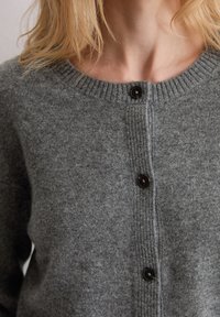 Cardigan en tricot gris avec un col côtelé et une fermeture à bouton. Il présente une texture douce et une coupe légèrement cintrée.