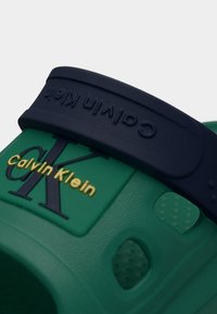 Πράσινο σανδάλι Calvin Klein με ρυθμιζόμενο λουράκι σε ναυτικό μπλε και ανάγλυφο λογότυπο της μάρκας με χρυσά γράμματα σε υφή επιφάνειας.