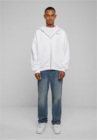 White zip-up hoodie with a front pocket, worn over a white t-shirt, paired with faded blue jeans and white sneakers. Minimalist design.