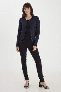 Cardigan marine avec un col festonné, fermeture à boutons sur le devant et manches longues, associé à un pantalon noir ajusté et des talons noirs.