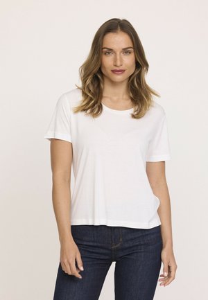 Femme avec des cheveux ondulés jusqu'aux épaules portant un t-shirt blanc à manches courtes et un jean bleu foncé, se tenant devant un fond clair.