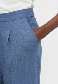 Χέρι με σκουρόχρωμο δέρμα που ακουμπά στην εμπρόσθια τσέπη μπλε υφασμάτινου, ψηλόμεσου παντελονιού, με λευκό τοπ.