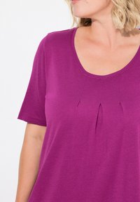 Top corto viola a maniche corte con scollo rotondo, caratterizzato da pieghe sotto lo scollo. Realizzato in un materiale morbido con una texture liscia.