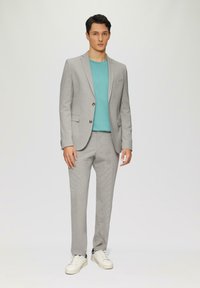 Completo grigio chiaro con due bottoni, abbinato a una camicia verde teal sotto. Sneakers bianche completano il look, caratterizzate da un tessuto liscio e una vestibilità su misura.