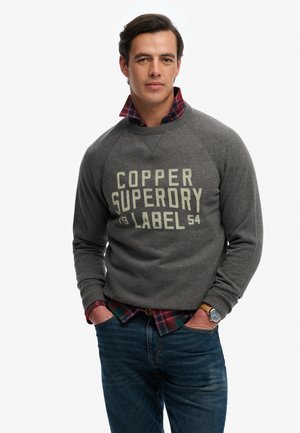Mand iført grå sweatshirt med teksten "Copper Superdry Label", rød ternet skjorte under, blå jeans og armbåndsur, hænderne i lommerne.