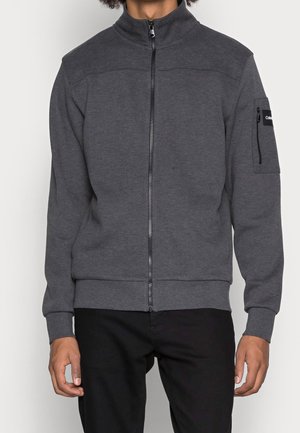 Mann trägt eine dunkelgraue Jacke mit Reißverschluss und hohem Kragen sowie einer Reißverschlusstasche am linken Ärmel, kombiniert mit schwarzen Hosen.