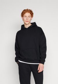 Sudadera negra de tela suave con bolsillo canguro, puños y dobladillo de canalé. Usada con una camiseta blanca debajo y pantalones de chándal negros.