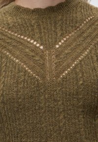 Pull en maille marron avec un motif ajouré formant un V près du col porté par une personne.
