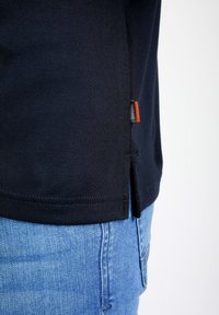 Navy bomba iz bombaž s stranskimi razporki, z majhno oznako z oranžnim poudarkom, v kombinaciji s svetlo modrimi jeans hlačami.