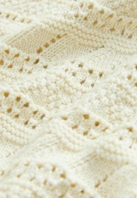 Tessuto lavorato a maglia di colore crema con un motivo testurizzato, caratterizzato da elementi a giorno e vari design dei punti. Aspetto morbido e accogliente.