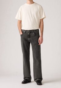 Homme portant un t-shirt blanc uni, un jean noir droit délavé, des chaussures noires et un bracelet argenté, debout avec les mains dans les poches.