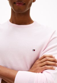 Personne portant un pull ras du cou rose clair avec un petit logo rouge, blanc et noir sur la poitrine, bras croisés devant.