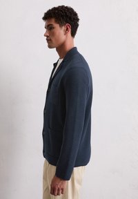 Marineblaue Strickjacke mit lockerer Passform, einer Brusttasche, fünf Knöpfen und einem Kragen, getragen über einem weißen Hemd.