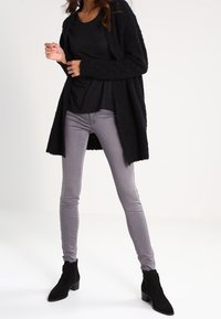 Cardigan negro de peluche sobre una camiseta negra, combinado con jeans ajustados gris claro y botines negros. Diseño sencillo con un ajuste relajado.