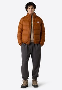 Brun pufferjakke med lynlås foran, som har en høj krave og quiltet design. Båret over en beige hættetrøje og mørkegrå sweatpants. Brune vandrestøvler.