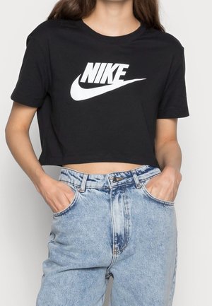 Osoba nosząca czarny krótki t-shirt Nike i jasnoniebieskie, wysokie spodnie jeansowe z rękami w kieszeniach na jednolitym tle.
