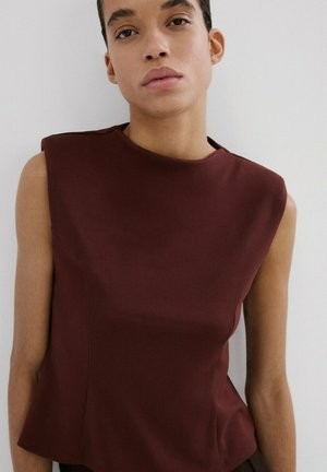 Donna con capelli corti e scuri che indossa una canotta marrone scuro, in piedi contro uno sfondo chiaro e uniforme.